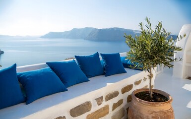 Fototapeta premium Blue pillows on white terrace wall overlooking sea, traditional Santorini island architecture with olive tree pot, luxury summer vacation resort view, greek mediterranean landscape.