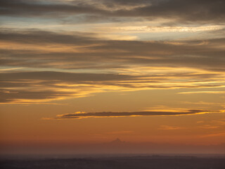 An evocative and dramatic image of a sunset over the Po Valley, where the sky is crossed by layered clouds in warm hues. The distinctive pyramidal silhouette of the Monviso mountain,