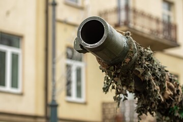 Close-up of a camouflaged tank cannon pointing towards the sky in an urban setting, with buildings, balconies and windows softly blurred in the background