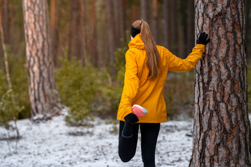 Fototapeta premium Woman stretching in a snowy forest while holding a pine tree, warming up before a winter run, focused and calm in cold natural surroundings.