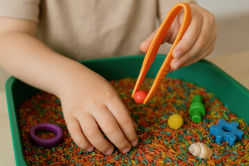 Child's Sensory Play: A child explores sensory learning through colorful rice, tongs, and small toys, fostering creativity and fine motor skills.