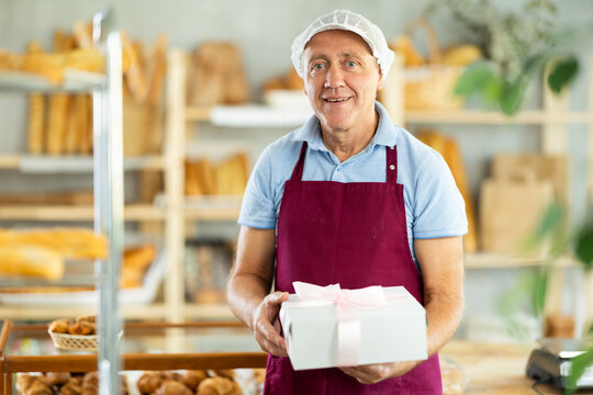 European senior man entrepreneur owner of bakery holds out cardboard box with pastries in hand, ready to meet buyer and sell goods. Seller presenting product in box with bow decoration