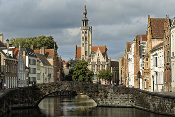 Obraz premium Historic canal scene in Bruges, Belgium, featuring the Poortersloge, a stone arch bridge and Gothic-style buildings with red-tiled roofs.