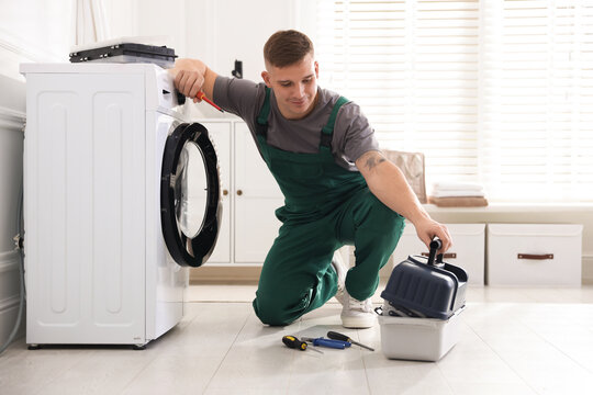 Repairman in uniform fixing broken washing machine with screwdriver and toolbox at home