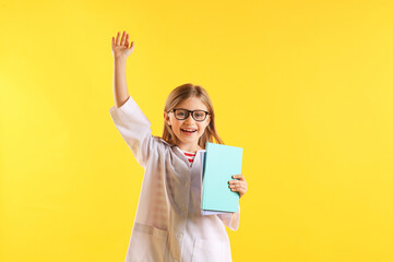 Portrait of smiling little girl in laboratory coat and glasses with book raising hand on yellow background. Child and science