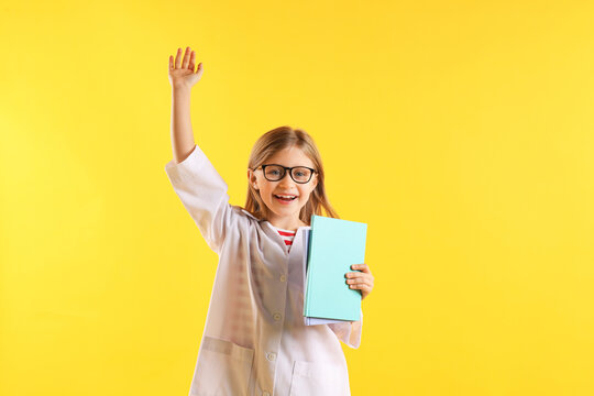 Portrait of smiling little girl in laboratory coat and glasses with book raising hand on yellow background. Child and science - Powered by Adobe