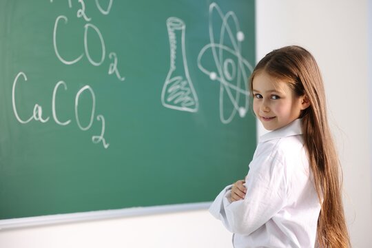 Smiling little girl in laboratory coat near green chalkboard with formulas indoors, space for text. Child and science