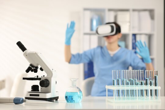 Modern technology. Laboratory glassware with samples, microscope on desk and doctor using virtual reality headset in office, selective focus