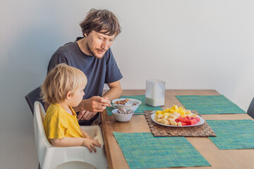 A father having breakfast at the table with his young son in a bright home kitchen, sharing a warm morning moment. Family bonding, parenting, everyday routine and healthy lifestyle concept