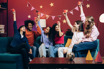 Diverse group of multinational friends celebrating joyful party at home sitting on sofa