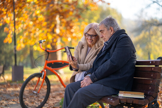 Senior couple enjoying a sunny autumn day in the park with a bicycle and phone - Powered by Adobe
