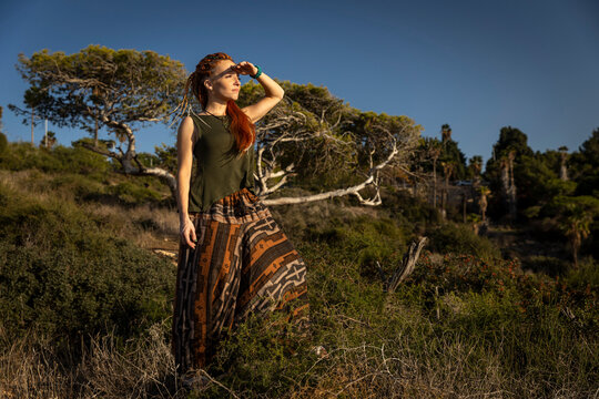 A focused woman with dreadlocks shades her eyes with her hand, gazing intently at the scenic, sunlit landscape from a grassy hillside.