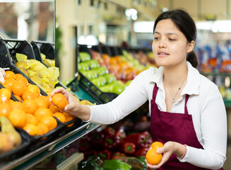 Young woman seller in apron lays out tangerines in supermarket