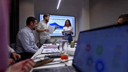 Diverse business professionals engage in a dynamic meeting, with a female presenter sharing data analysis results on a large screen. - Powered by Adobe