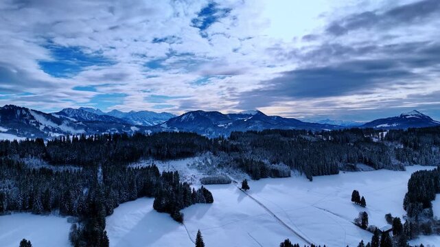 Lonely car rides through the snow-covered valley near the pine-tree woods. Beautiful mountains at backdrop. Clouds cover the sky above. Aerial view.