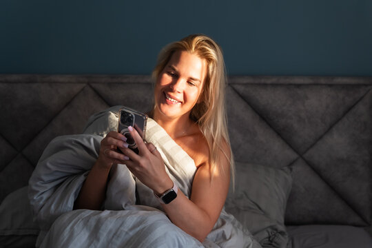 Happy blonde woman sitting in bed using a smartphone and smiling while checking social media in the morning sunlight