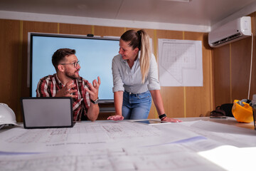 Two architects are discussing a blueprint in their office container on a construction site, analyzing the project details and collaborating on the design