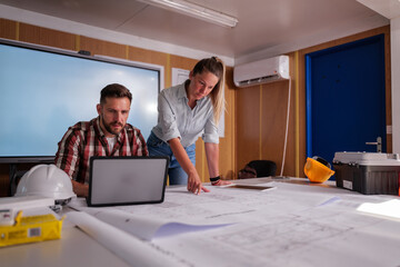 Two engineers are examining blueprints on a table in their office at a construction site, using a laptop and tablet to aid their analysis and planning