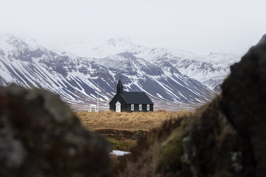 View of stark black church atop golden fields contrasted against snow-dusted mountains under a grey sky, Snaefellsnes, Eyja- og Miklaholtshreppur, Iceland.