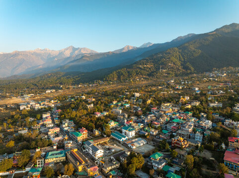 Aerial view of a settlement nestled amidst lush trees, with the majestic Dhauladhar range rising in the backdrop, Bir, Himachal Pradesh, India.