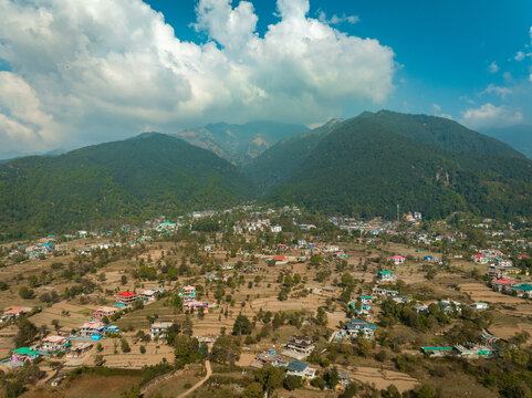 Aerial view of the village nestled among rolling hills and verdant forests, with a bright blue sky and fluffy white clouds above, Bir, Himachal Pradesh, India. - Powered by Adobe
