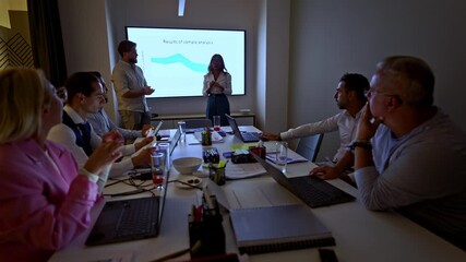 A business team engages in a productive meeting, discussing data analysis and strategic planning. Colleagues gather around a table with laptops, while a presenter shares insights on a large screen. - Powered by Adobe