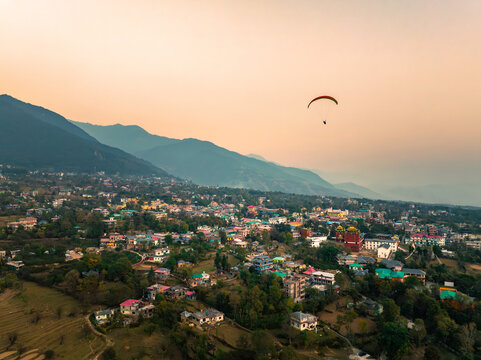 Aerial view of a lone paraglider soaring over the town nestled amidst rolling hills, bathed in the warm glow of the setting sun, Baijnath, Himachal Pradesh, India.