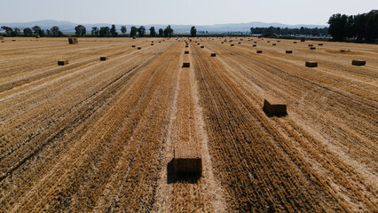 Wide aerial shot of a large, harvested grain field with straw bales and linear patterns, leading towards a distant town and mountains under a clear sky