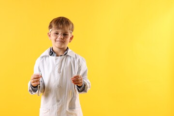Smiling little boy in laboratory coat and protective goggles with test tubes on yellow background,...