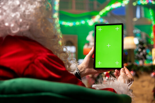 Close up of Santa Claus using mockup tablet to monitor toy shipments during December festive season. Man playing Christmas character with white beard follows gift deliveries with green screen device