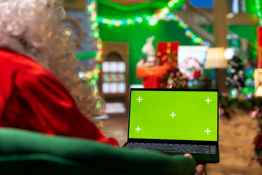 Santa Claus using mockup laptop in festive ornate workshop to browse online store, purchasing sleighs. Man surrounded by Christmas lights decorations using internet on green screen notebook