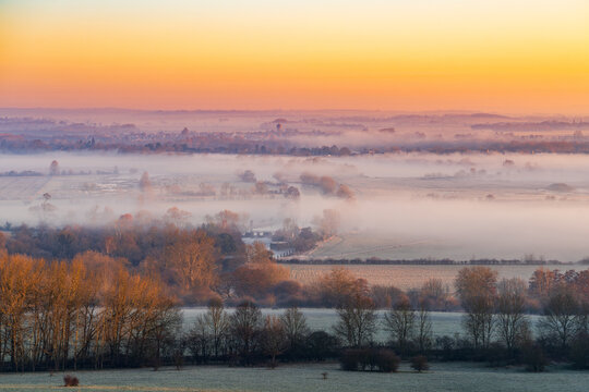 View of a serene landscape where golden sunlight kisses the frosty ground and veils the trees in a gentle, ethereal mist, The Chilterns, Oxfordshire, England, United Kingdom.