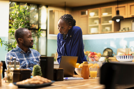 African American freelancer sitting at a kitchen table with a laptop, sending emails and explaining business tasks to his girlfriend. Couple enjoys a relaxed home lifestyle with coffee.