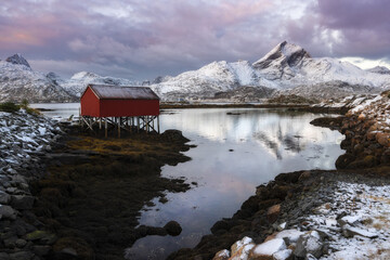 View of a vibrant red boathouse perched on stilts amidst the tranquil waters, mirroring the snow-capped mountains, in Reine, Nordland, Norway.