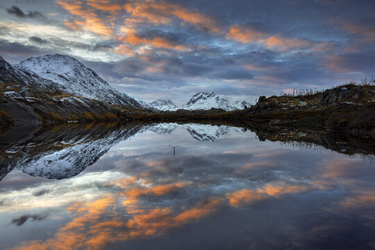 View of snow-dusted mountains mirrored in the tranquil lake under a vibrant sky painted with fiery orange and blue hues, Reine, Nordland, Norway.