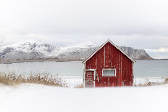 View of a rustic red cabin nestled in the snow against the backdrop of stark mountains and tranquil waters, Reine, Nordland, Norway.