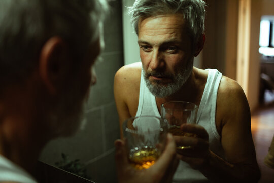 Mature man looking pensive with whiskey at bathroom mirror