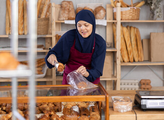 Saleswoman in Muslim clothes stands behind a counter with scales and chooses a croissant. Woman in...