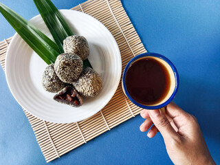 Indonesian sesame balls known as onde onde with pandan accents on a white plate, one bitten to reveal red bean center, paired with a hand holding a cup of tea in soft bright lighting.