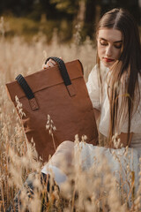 Natural Beauty: A woman in a white summer dress sitting on a golden grain field, holding a large, brown, ecological bag against the background of soft light.
