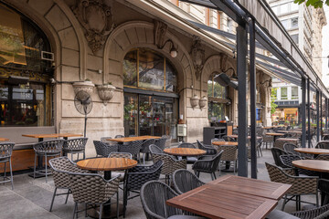 Outdoor cafe terrace on a city street with tables, chairs, greenery, and large trees providing shade