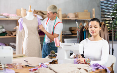 Attentive woman tailor sewing textile using sewing machine and young male assistant working with fabric and mannequin in small fashionable sewing atelier