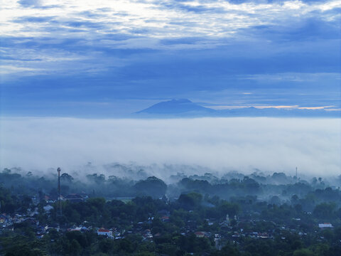 Aerial view of a mist-shrouded village, with mountain peaks piercing the mist under a cloudy sky, Klaten Regency, Central Java, Indonesia.
