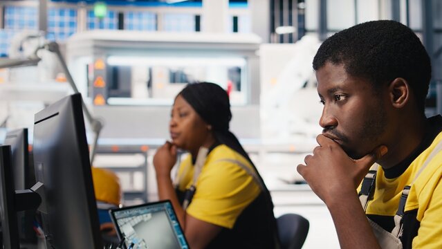 Male specialist adjusts machine parameters for solar panel production, working at a high tech manufacturing plant. Black engineer analyzing technical data on a computer. Camera B.