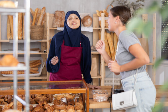 Smiling Arab woman in traditional Muslim attire and apron standing at counter in warm and inviting bakery filled with freshly baked goods, sharing friendly conversation with young female customer