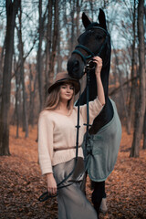 Woman stands beside horse in forest during fall season while wearing a hat and sweater © Тамара Печеная