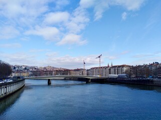 Naklejka premium A bridge crosses the Saône River with the backdrop of Lyon's hill and historic skyline, featuring modern construction with cranes towering over new white buildings.