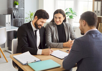 Smiling adult couple sitting at desk with male agent and signing documents in office. Happy man and woman making purchase and closing successful deal with real estate agent or financial advisor.