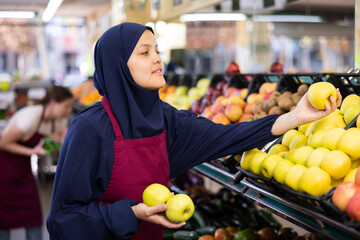 Young woman seller in hijb and apron sells apples in grocery store