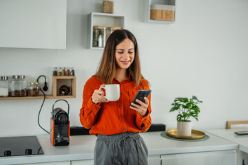 Woman drinking coffee browsing smart phone in modern kitchen